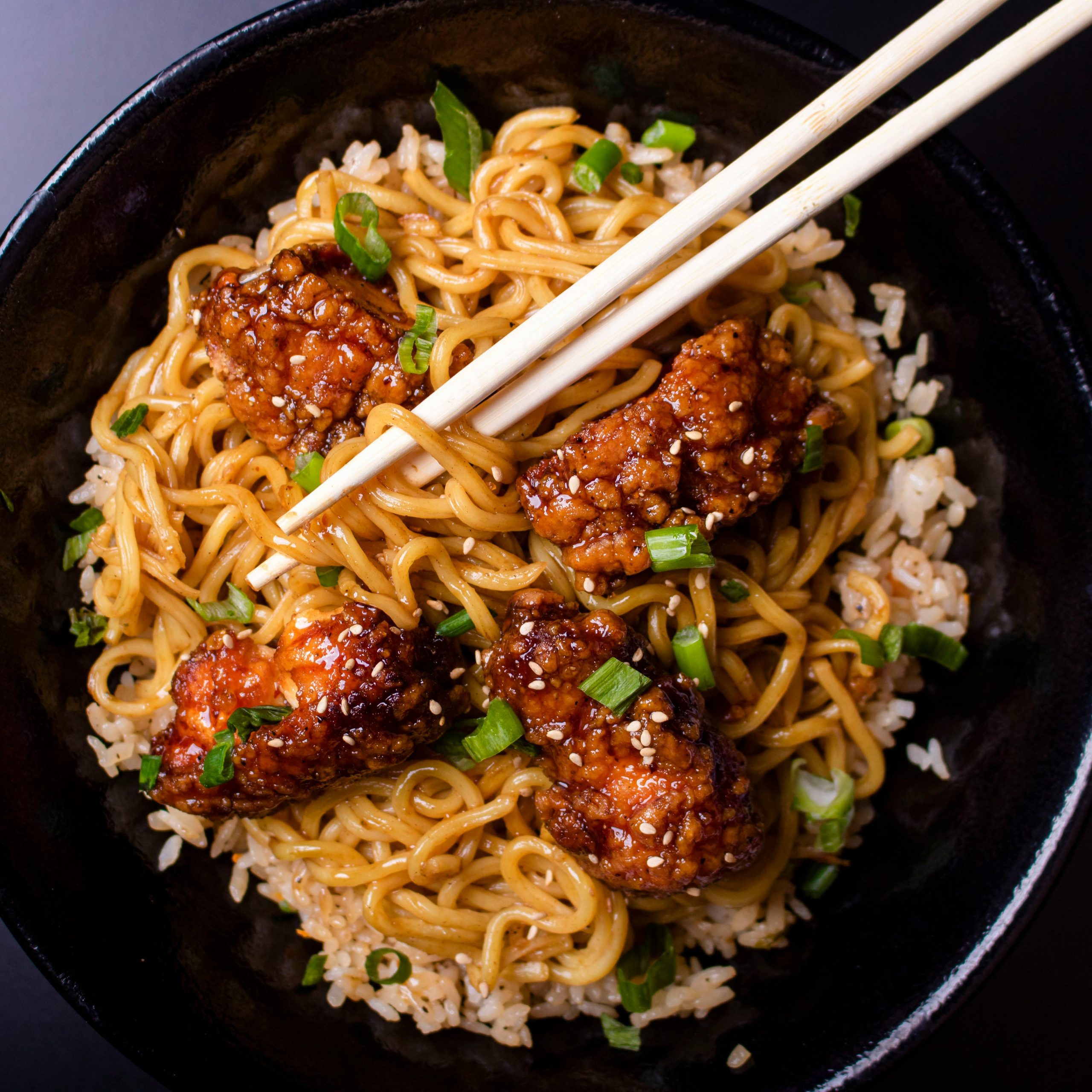 Top view of sesame chicken noodles garnished with green onions in a black bowl.